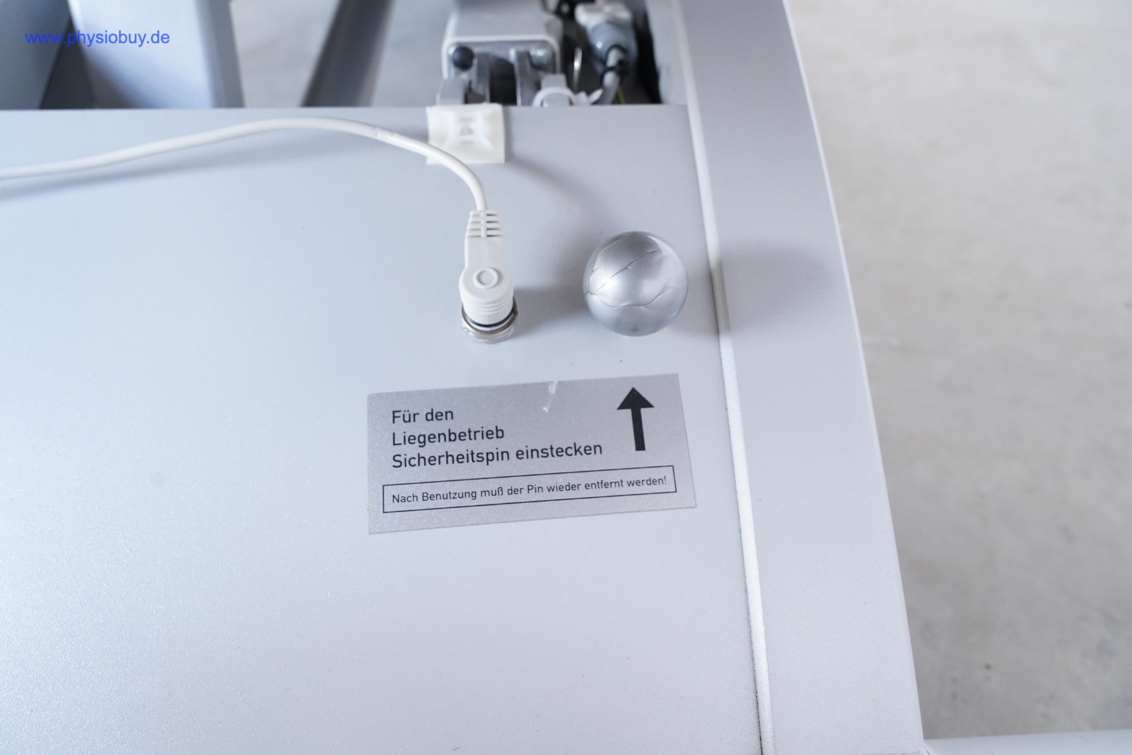 FREI THERA Standard Therapieliege: Hinweisschild zum Sicherheitspin neben Kabelstecker und Metallkugelknauf
