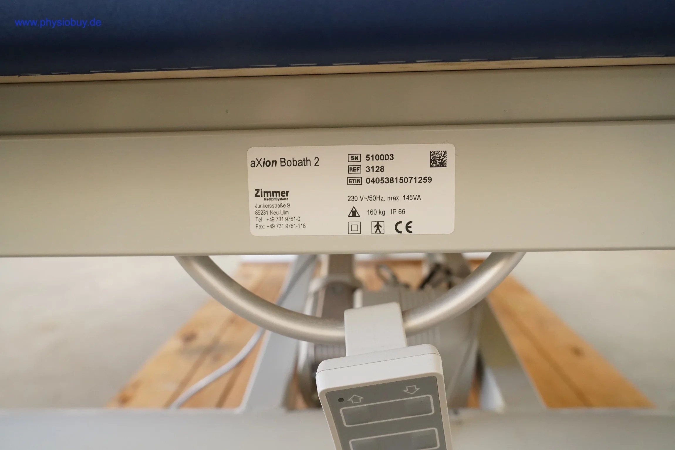 Typenschild der Zimmer aXion Bobath 2 am Rahmen, darunter Ring-Fußbügel und Handschalter, im Hintergrund Holzpaletten