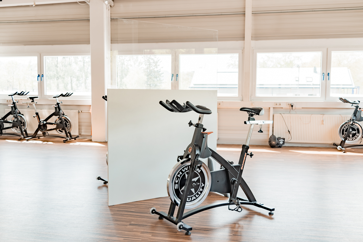 Trennwand Standard mit hellem Unterteil steht neben einem Indoor-Cycle vor Fensterfront