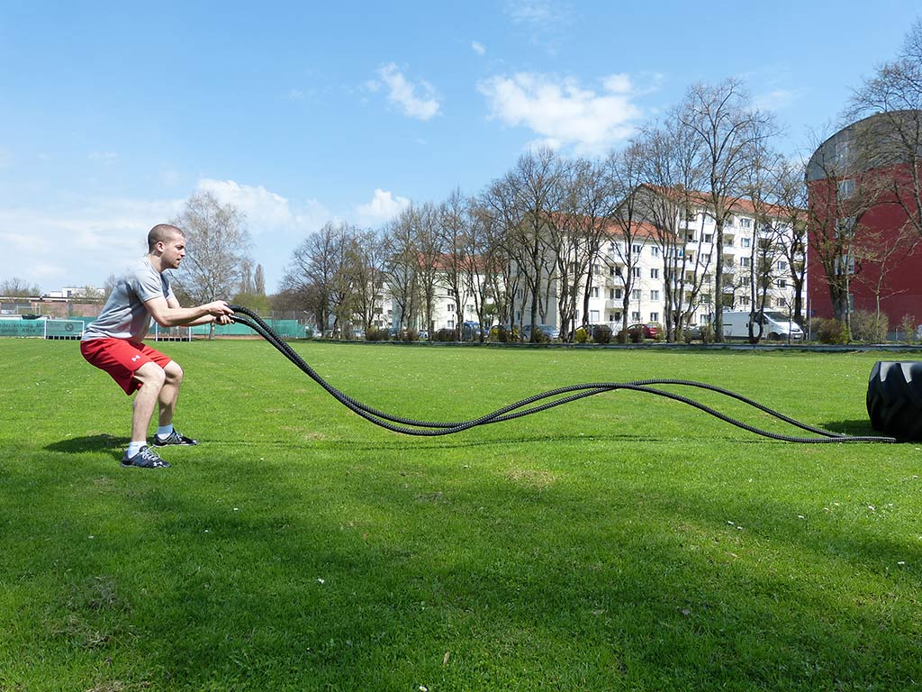 Mann zieht im Ausfallschritt ein langes schwarzes Battle Rope über eine Wiese, Wohnhäuser im Hintergrund