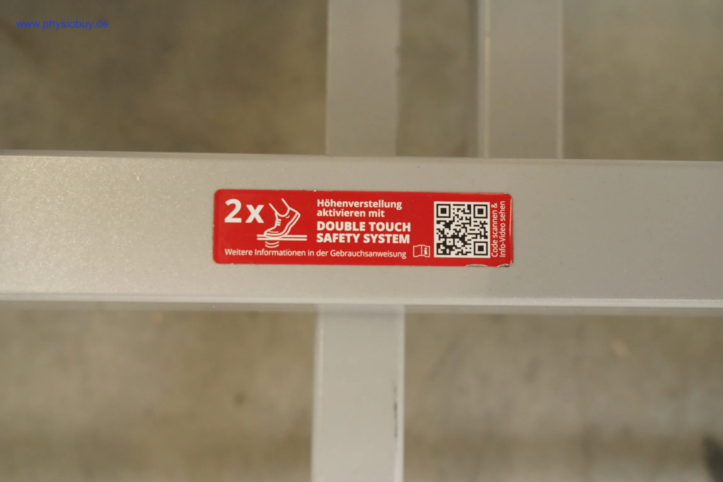 Pino XPert+ Therapieliege: roter Aufkleber Double Touch Safety System mit QR-Code auf grauem Metallrahmen