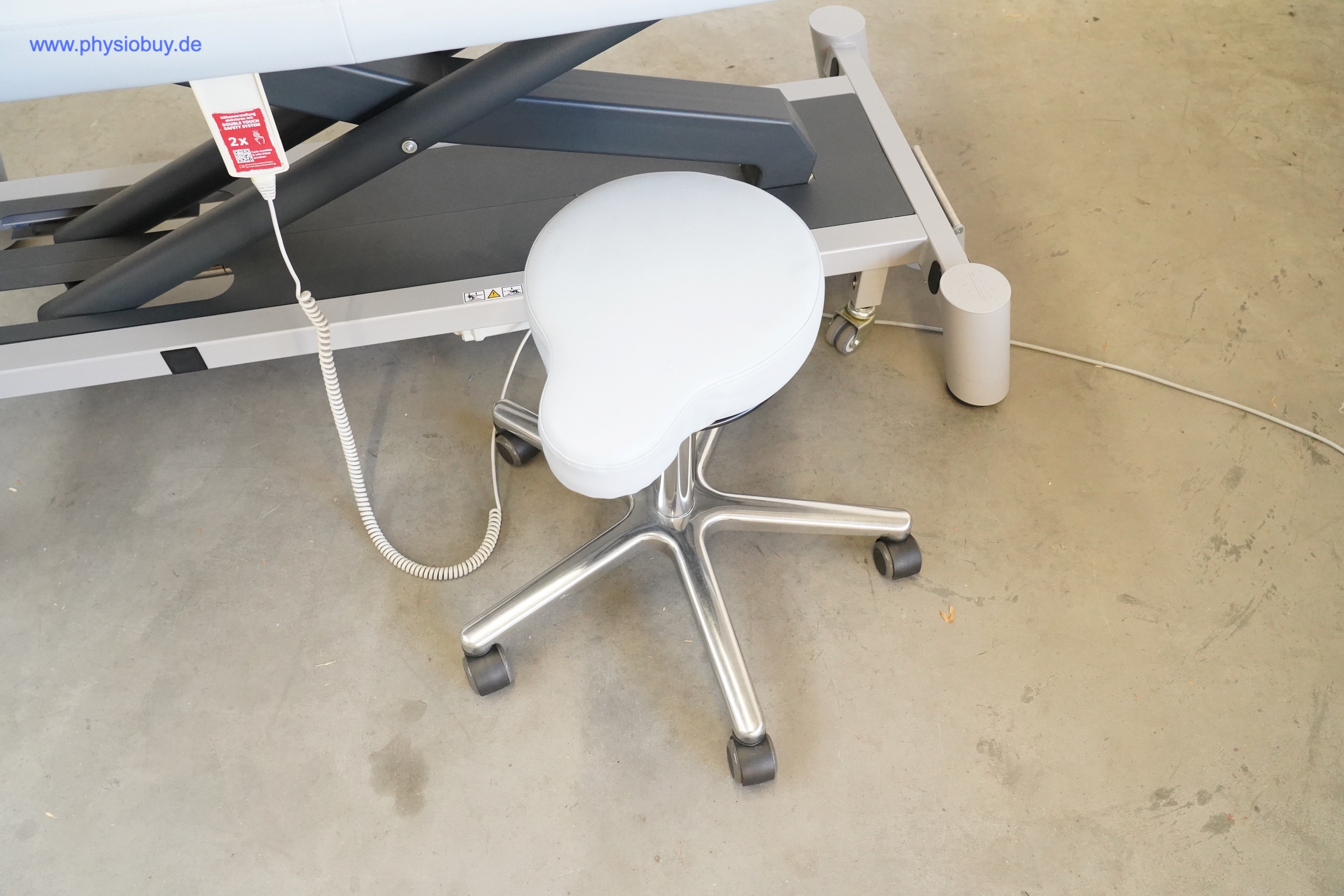 Heller Rollhocker mit Fünfsternfuß vor der Pino XPert+ Therapieliege, links Handschalter mit Spiralkabel, Betonboden