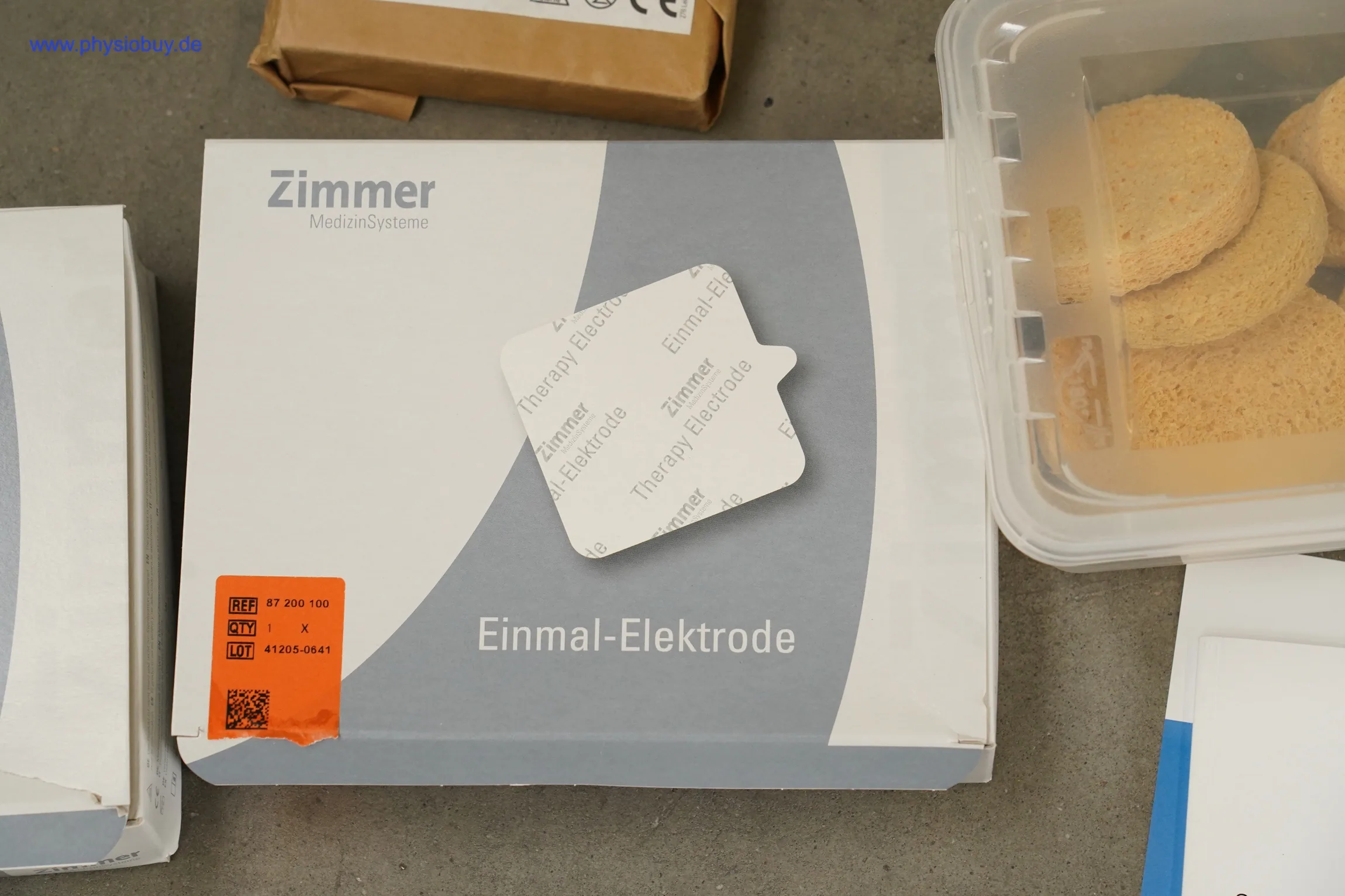 Verpackung der Zimmer Einmal-Elektrode mit aufgedrucktem Elektrodenpad, daneben Schwammelektroden in einer Plastikbox