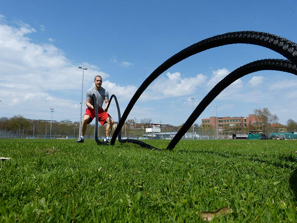 Mann kniet im Ausfallschritt auf einer Wiese und schwingt ein schwarzes Battle Rope wellenförmig