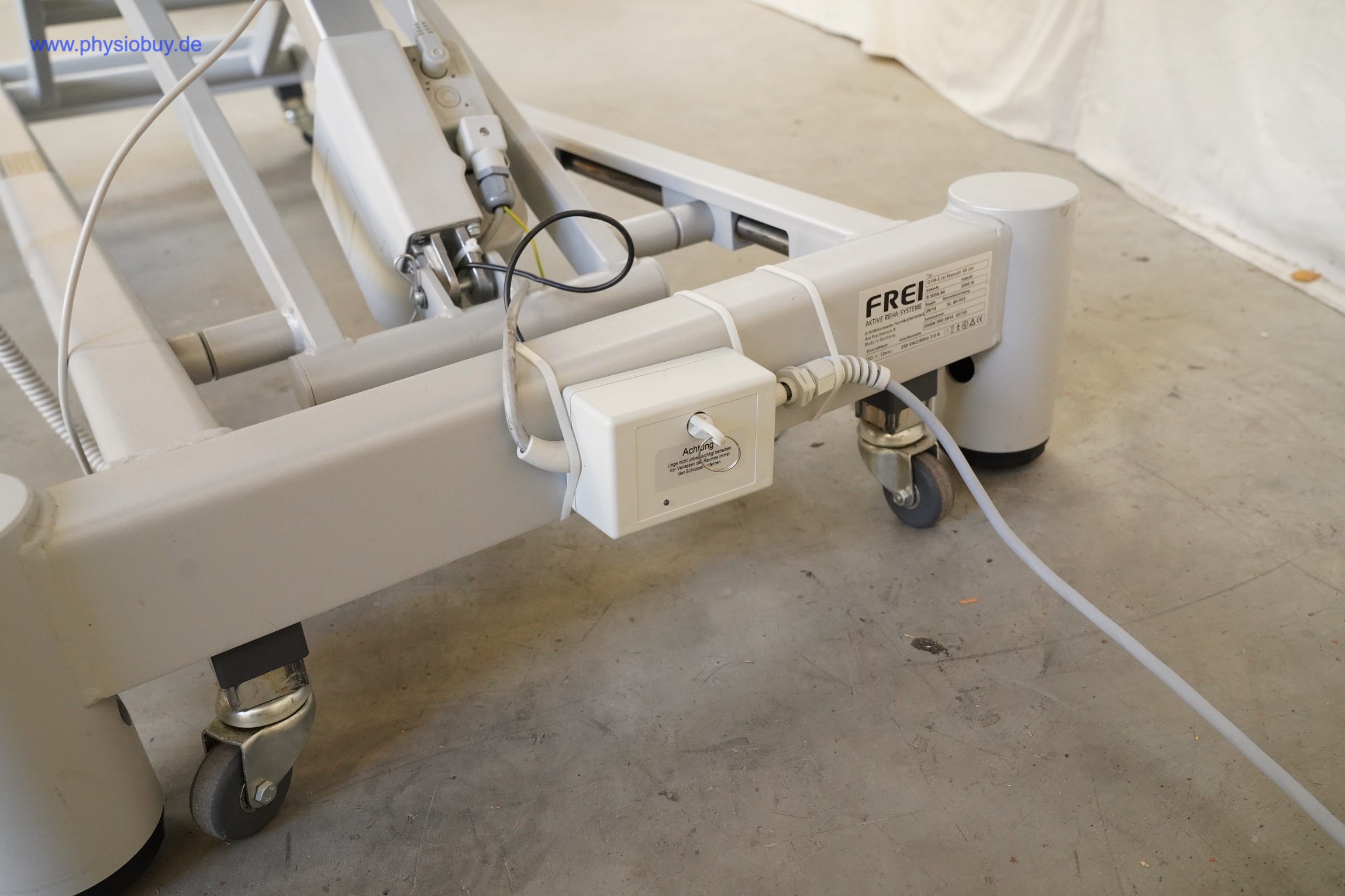 FREI Gym-X (e) Therapieliege, Detail des Untergestells mit Rollen, Typenschild, Steuerbox und Netzkabel auf Hallenboden