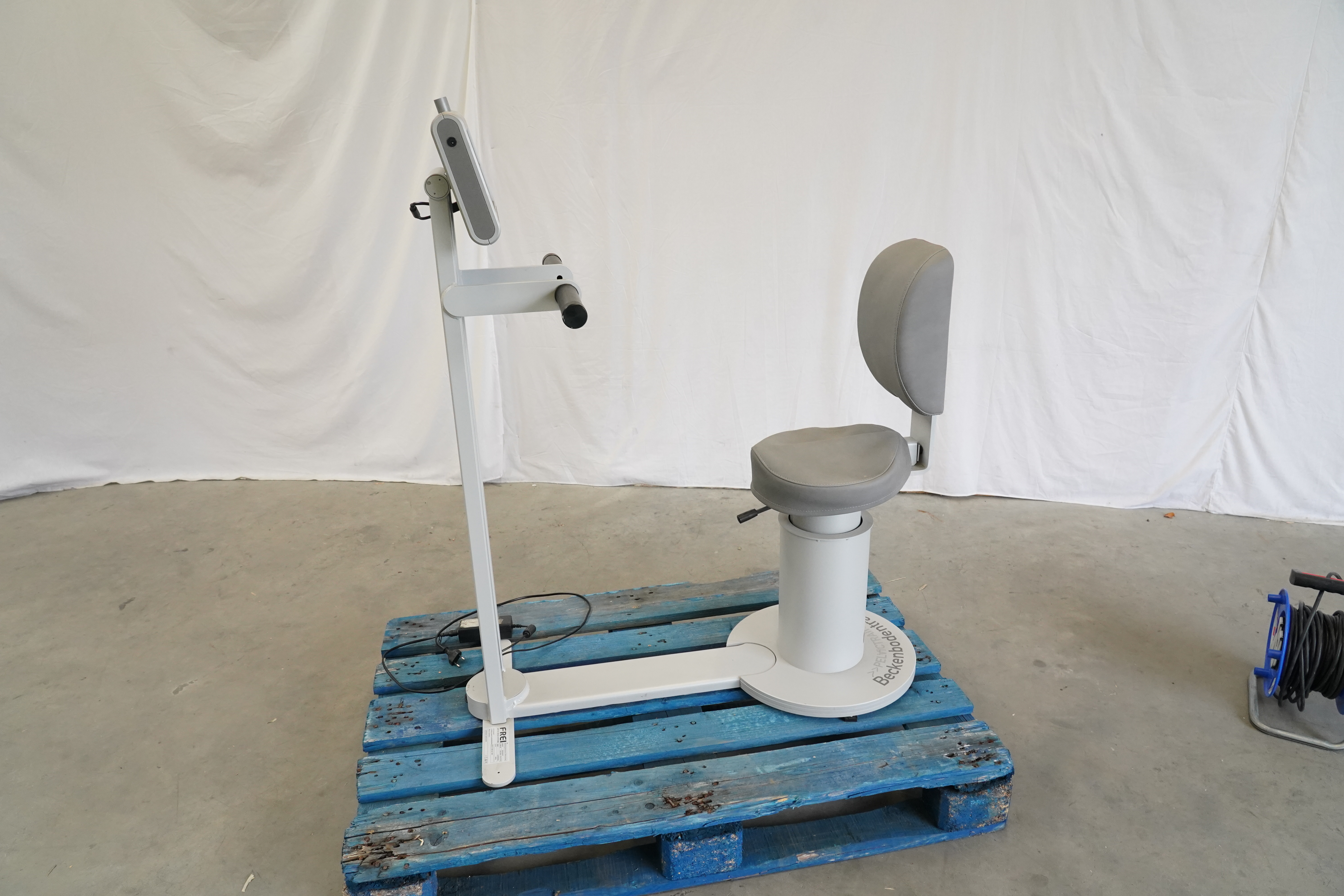 FREI Beckenboden/Pelvictrainer mit grauem Sitz und Rückenlehne, seitlich auf einer blauen Palette vor weißem Vorhang