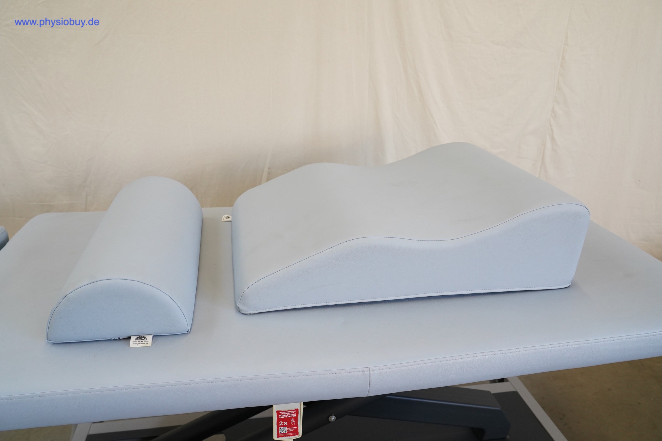 Halbrolle und Beinlagerungskissen in Hellblau auf der Polsterfläche der PINO XPert+ Therapieliege, vor heller Wand