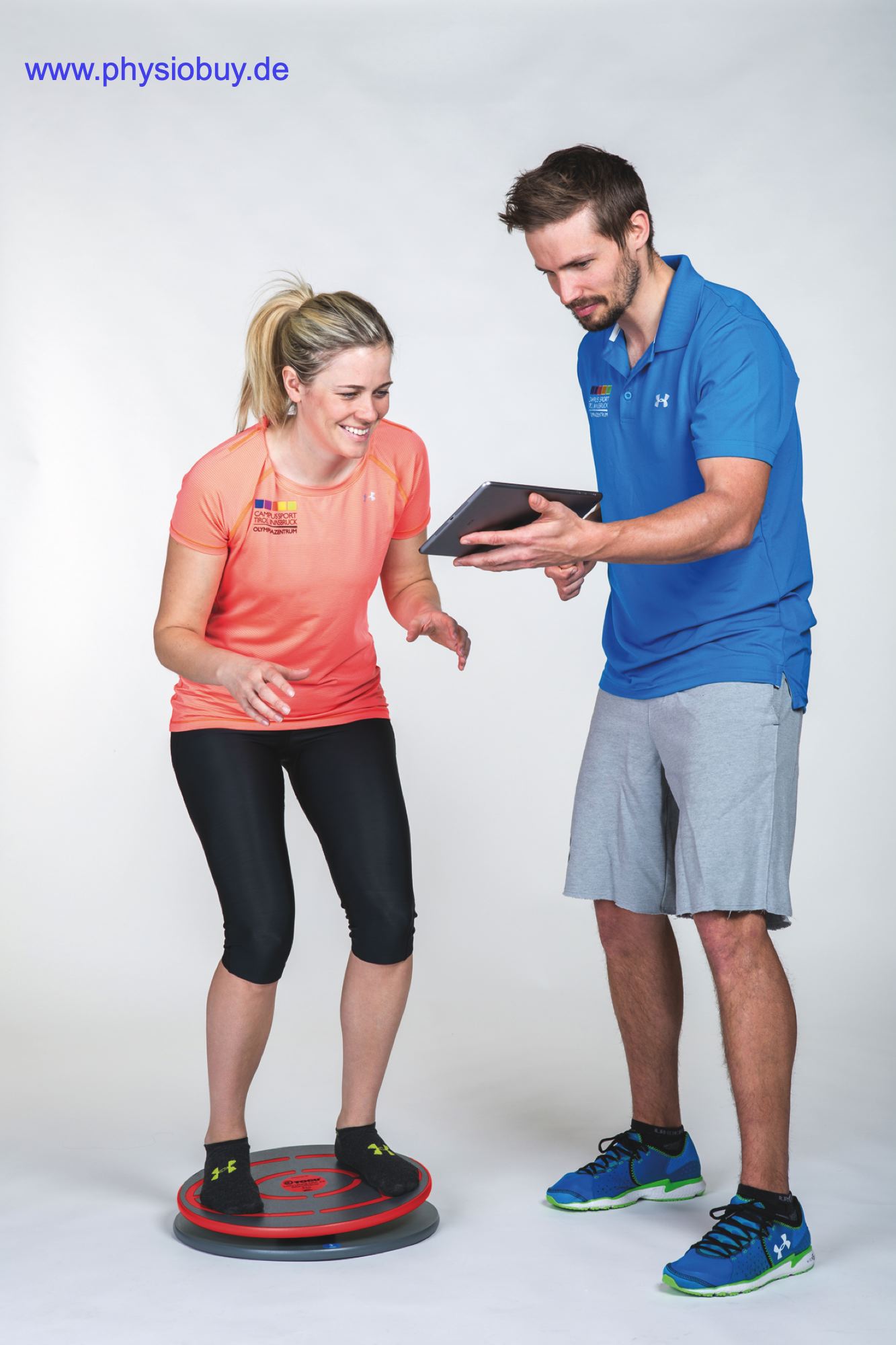 Frau balanciert auf der Challenge Disc, Trainer zeigt die zugehörige App auf einem Tablet