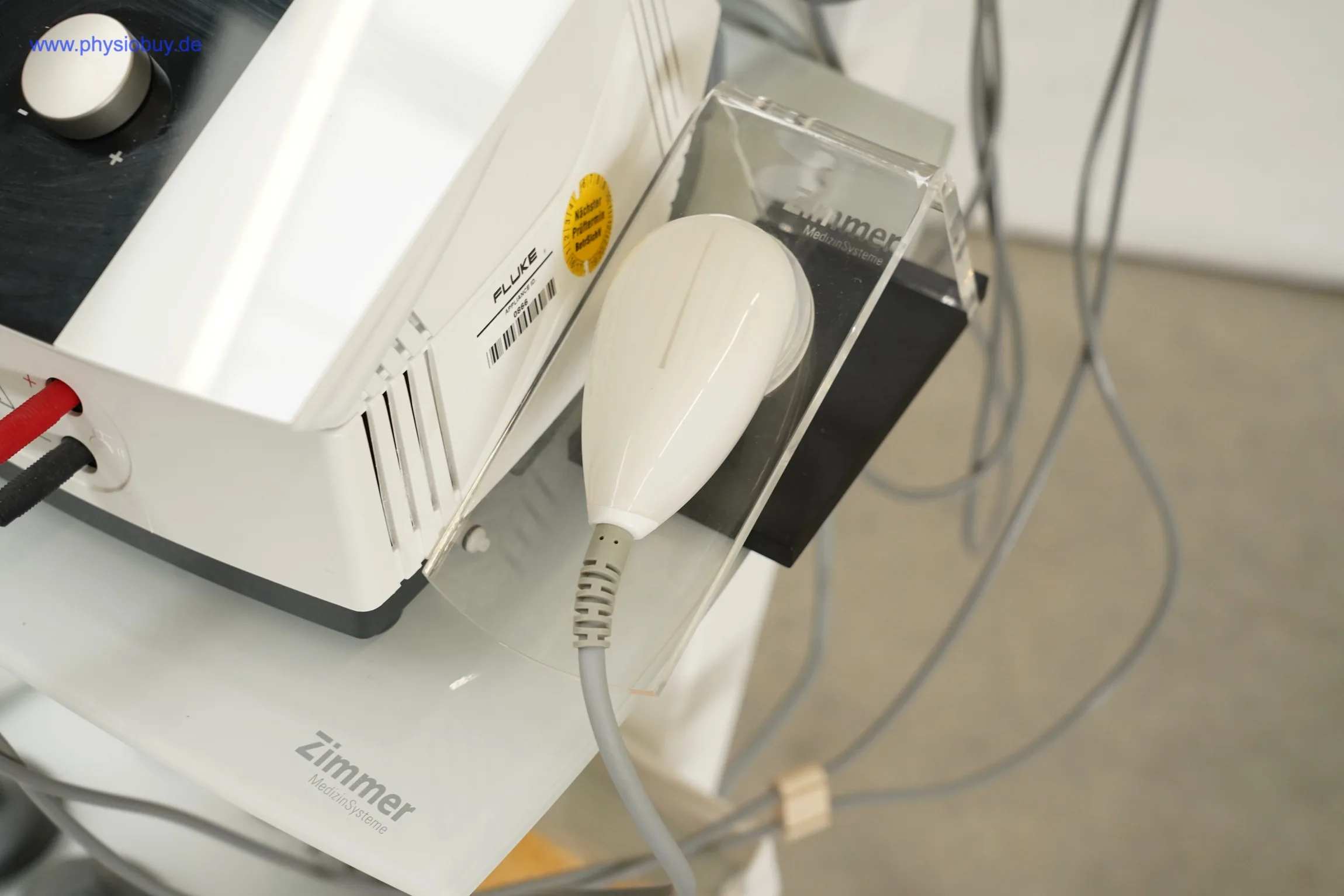 Zimmer Soleo SonoStim: Ultraschall-Applikator in transparenter Halterung neben dem Gerätegehäuse, Kabel hängen herab