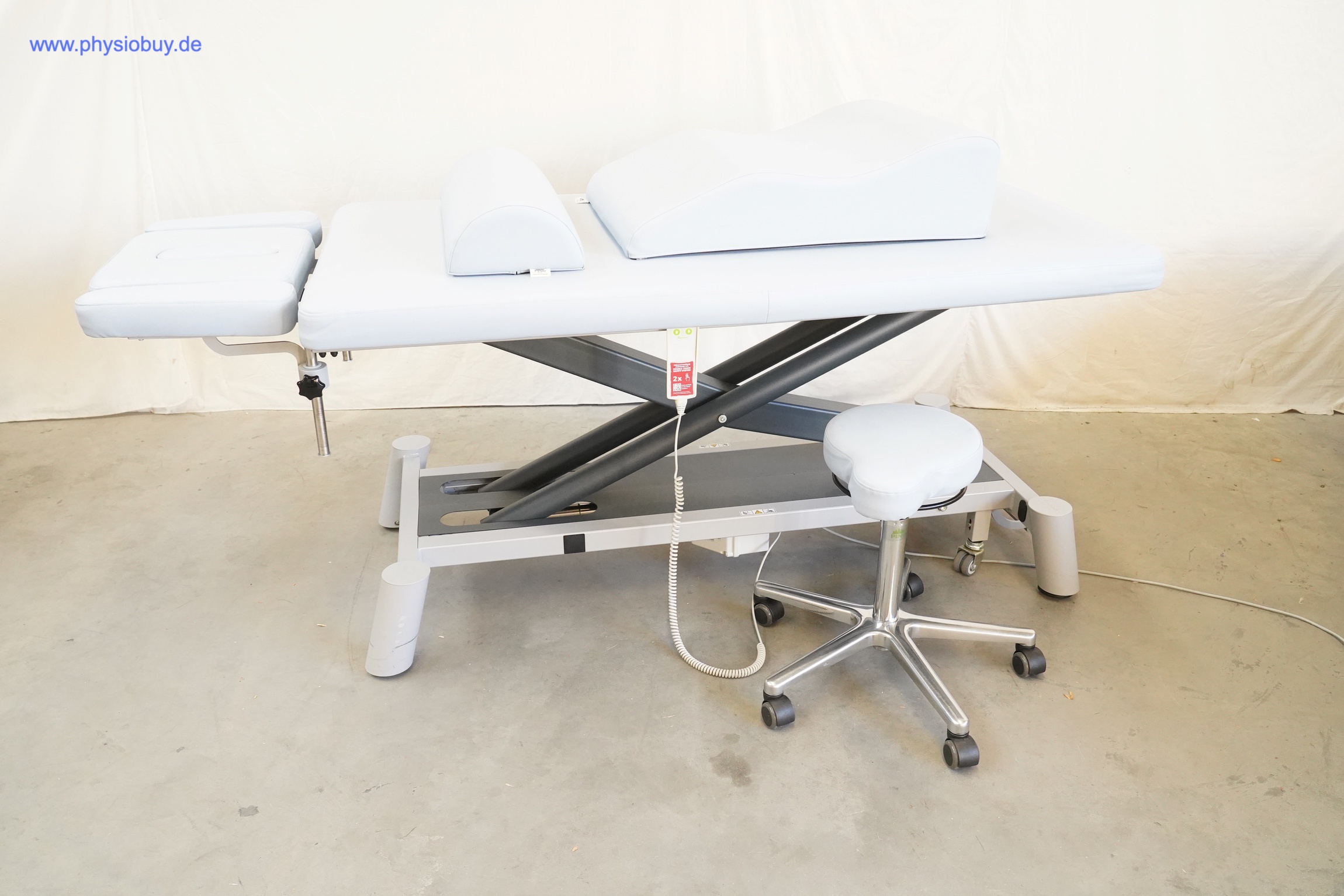 PINO XPert+ Therapieliege seitlich mit Scherenhubgestell, Lagerungskeilen, Handschalter und einem Rollhocker davor
